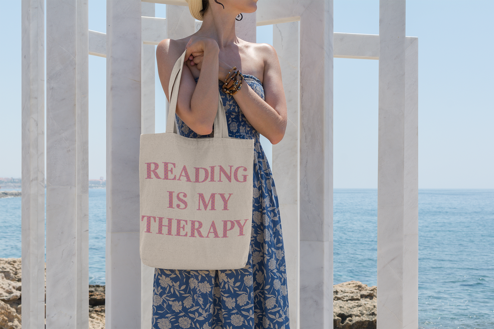 Woman holding a canvas bag which reads, Reading Is My Therapy. She is standing by the beach.
