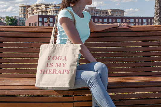 Woman sitting on a park bench with a canvas bag which reads, Yoga Is My Therapy.